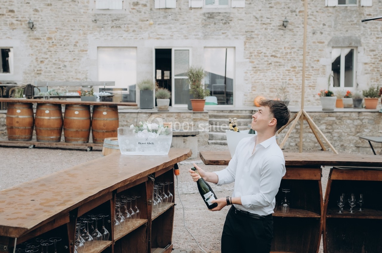 Un homme ouvre une bouteille de champagne derrière un bar extérieur, devant un bâtiment en pierre.