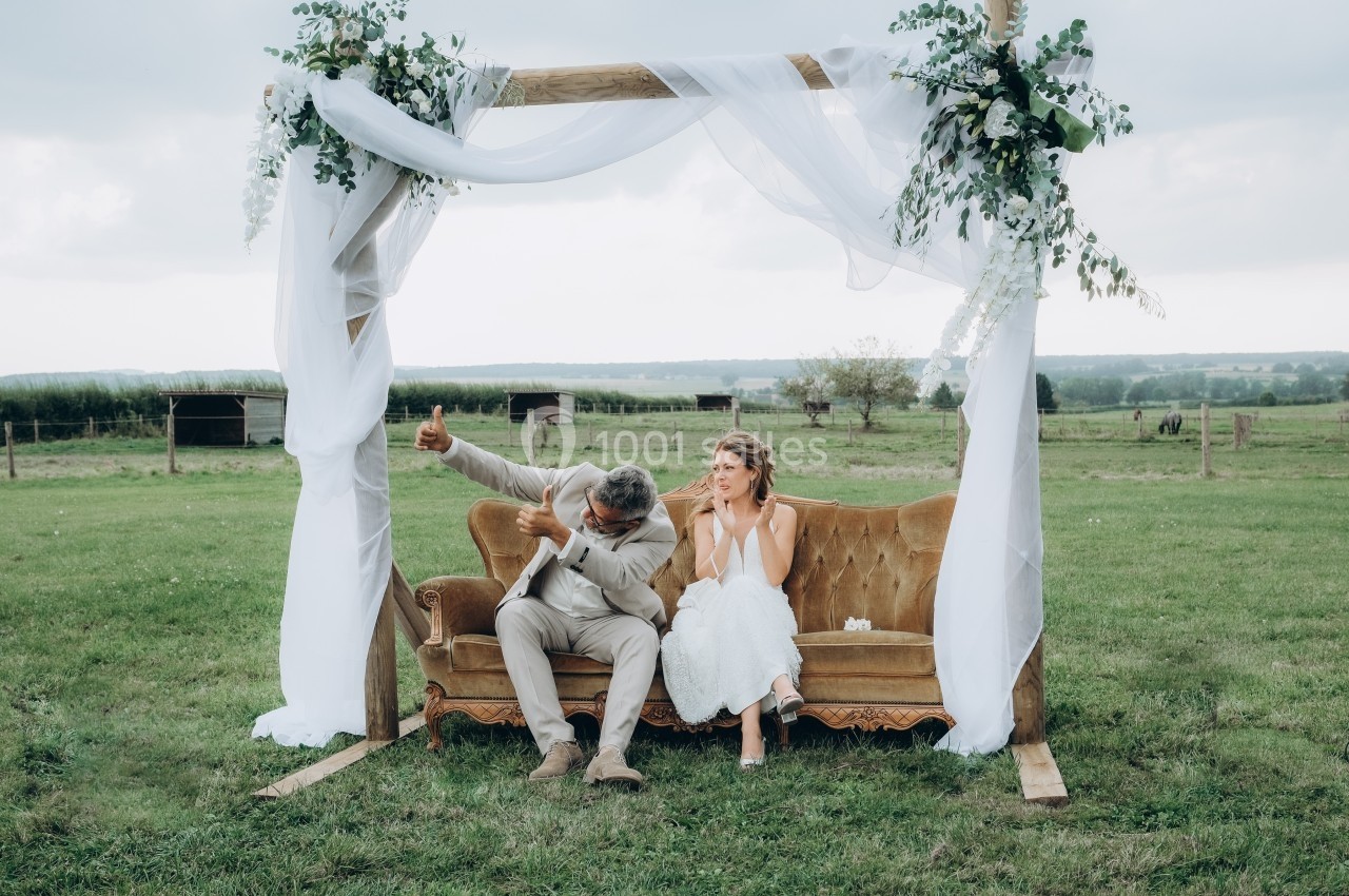 Un couple assis sur un canapé en plein air sous une arche décorée de voilages et de fleurs, dans un champ.