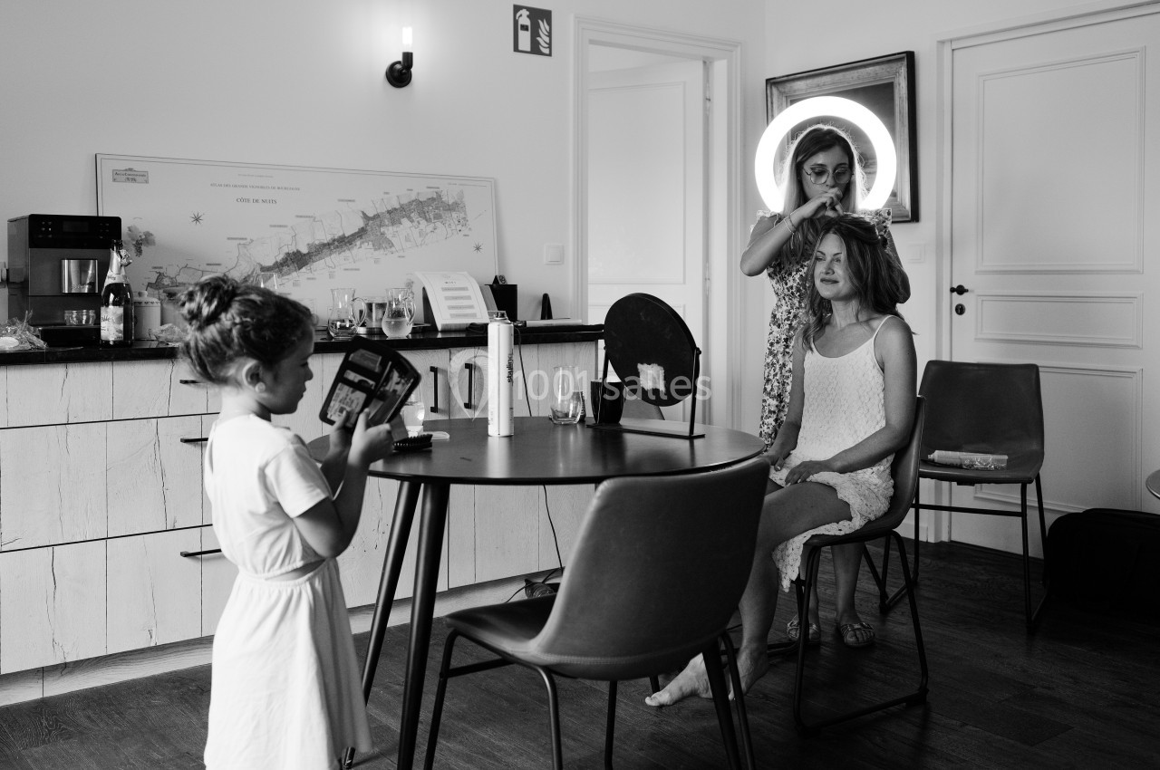 Une femme se fait coiffer dans une pièce tandis qu'une enfant regarde un livre près d'une table.