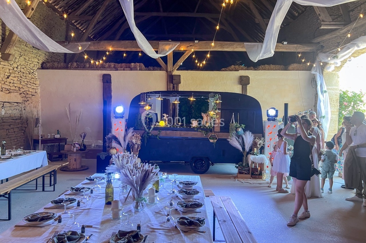 Salle de réception décorée pour un événement, avec tables dressées, guirlandes lumineuses et un bar mobile en arrière-plan.