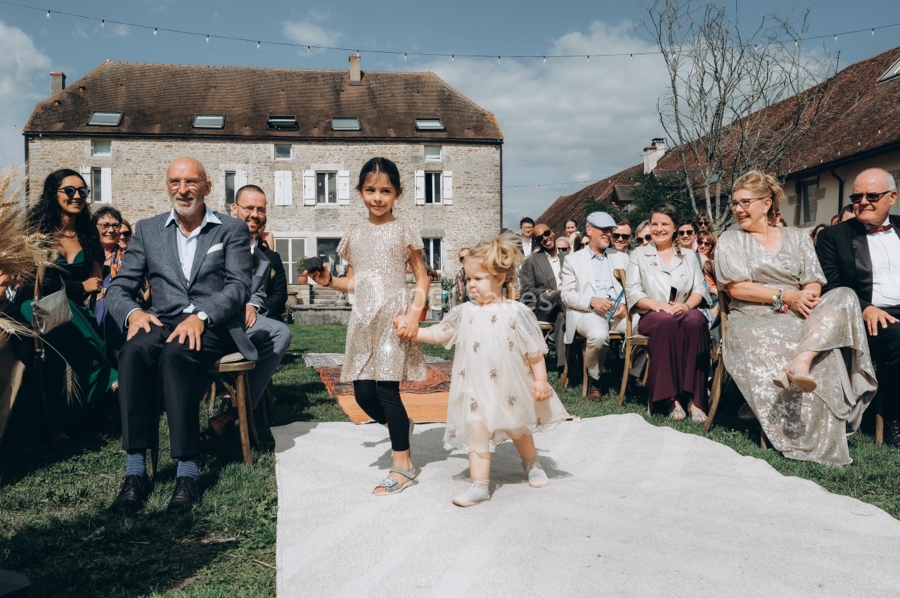 Deux enfants marchent main dans la main sur une allée blanche lors d'une cérémonie en extérieur, entourés d'invités…