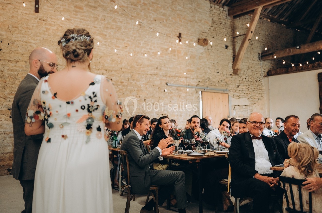 Un couple en tenue de mariage s'adresse à des invités assis dans une salle en pierre décorée de guirlandes lumineuses.