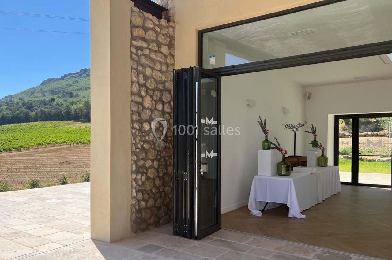 Entrée d'une salle lumineuse avec table décorée, ouverte sur une terrasse en pierre et un paysage de vignobles.