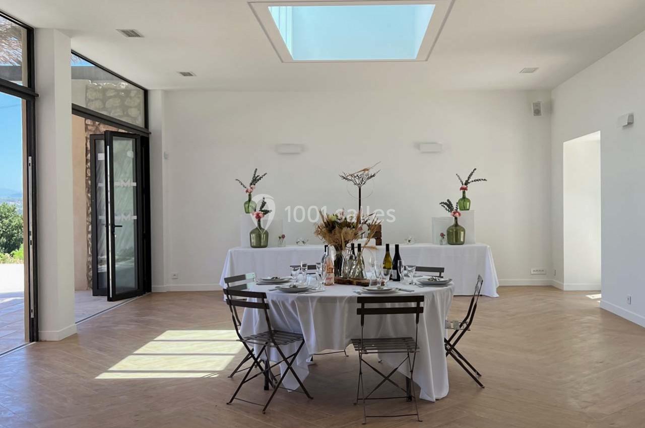 Salle lumineuse avec une table dressée pour un repas, décorée de fleurs séchées, et des baies vitrées ouvertes sur l…