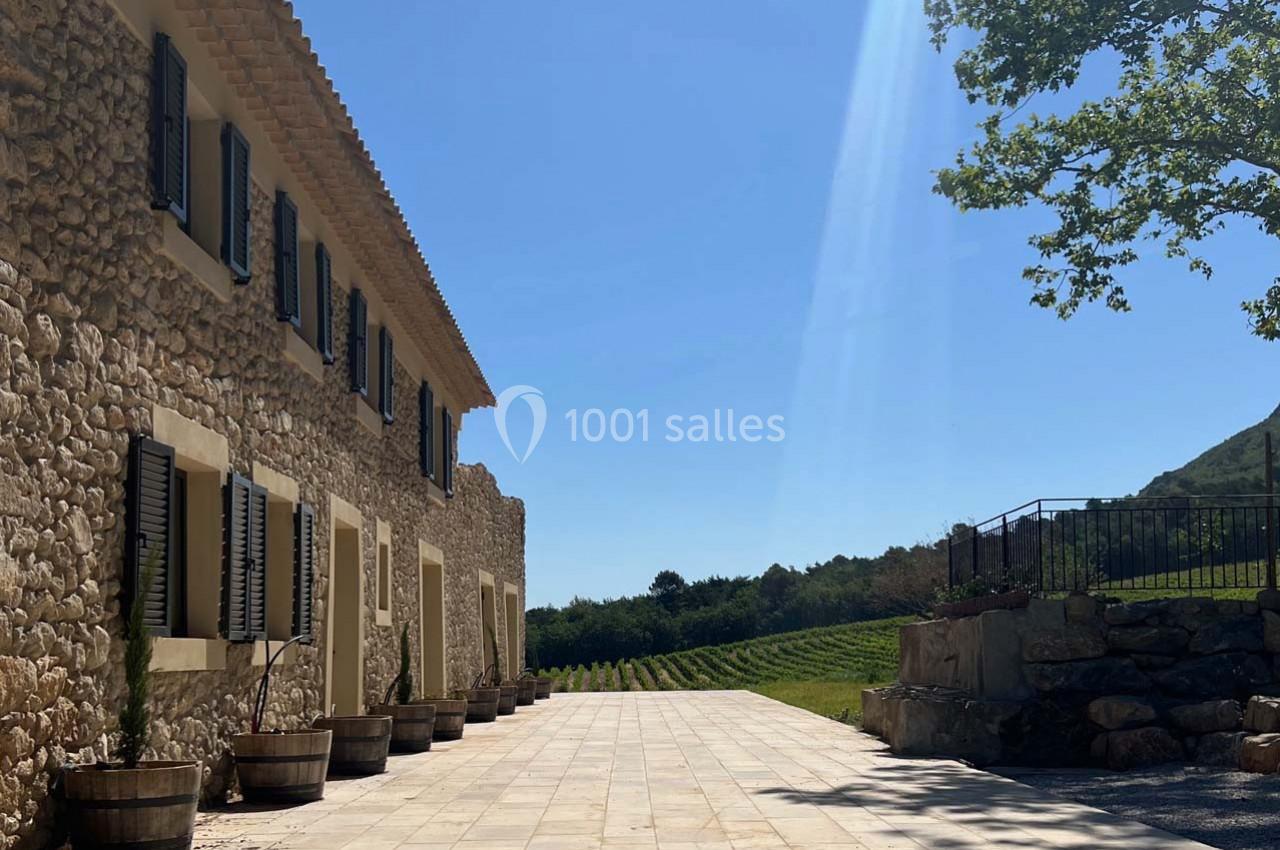 Façade en pierre d'une maison avec volets noirs, terrasse pavée et vignoble en arrière-plan sous un ciel bleu.
