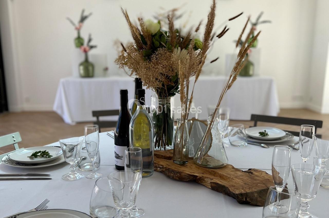 Table dressée avec nappes blanches, décorations florales séchées, bouteilles de vin et vaisselle élégante.