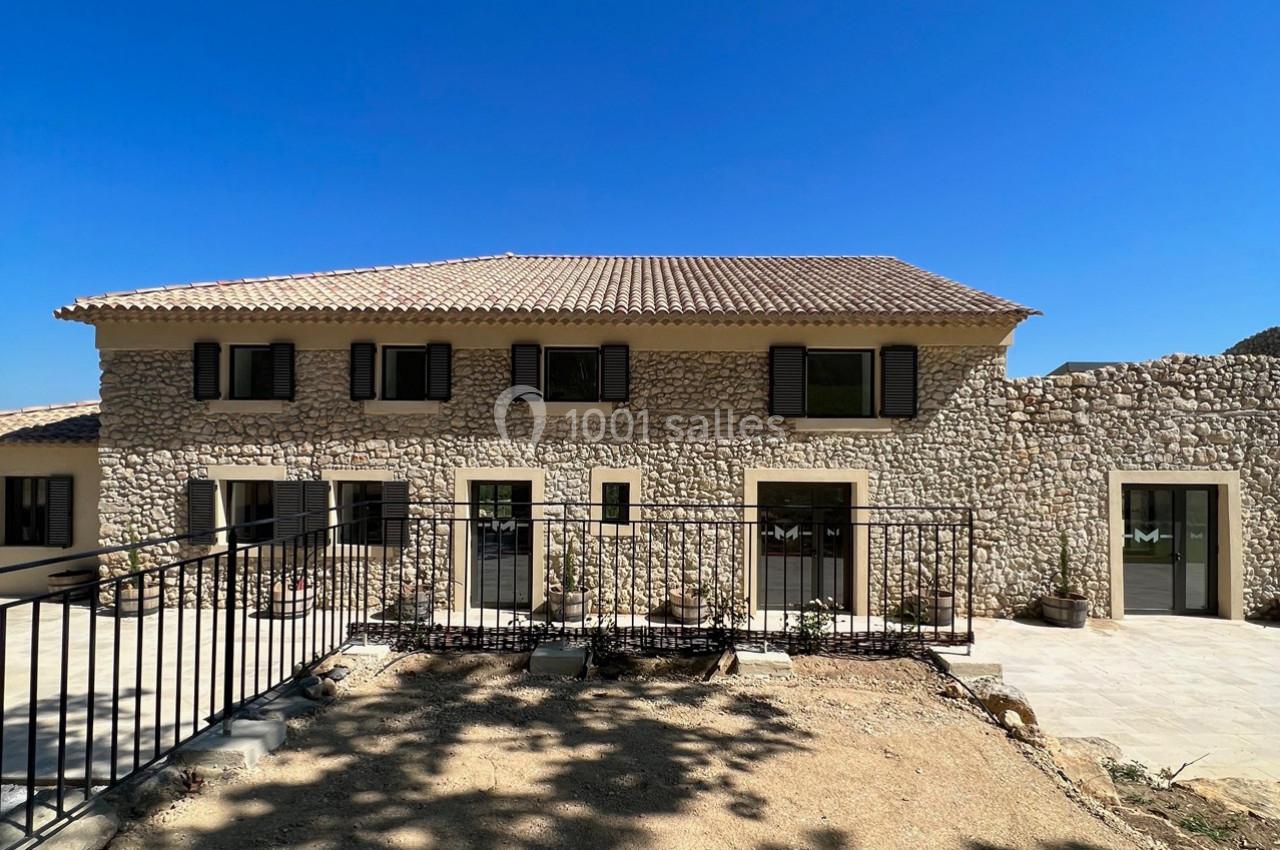Façade d'une maison en pierre avec volets noirs, terrasse ensoleillée et sol en terre devant une clôture métallique.