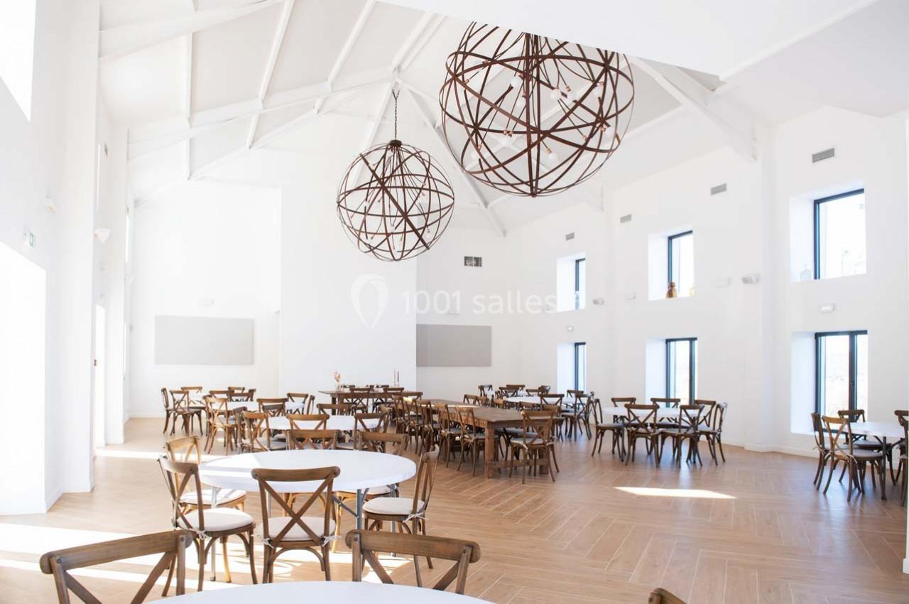 Salle lumineuse avec de grandes fenêtres, des tables rondes et des chaises en bois, décorée de suspensions métalliques.