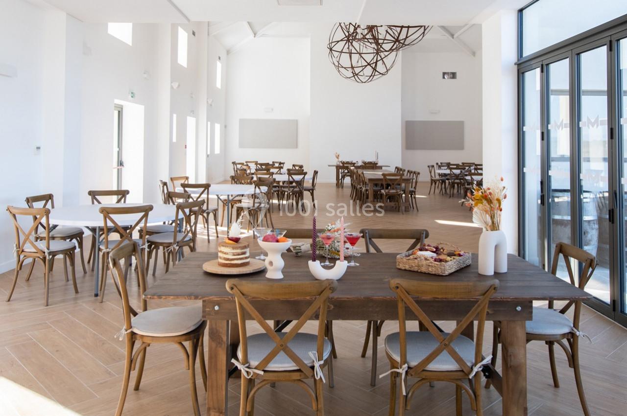 Salle lumineuse avec tables en bois, chaises croisées et décorations minimalistes, vue sur de grandes baies vitrées.