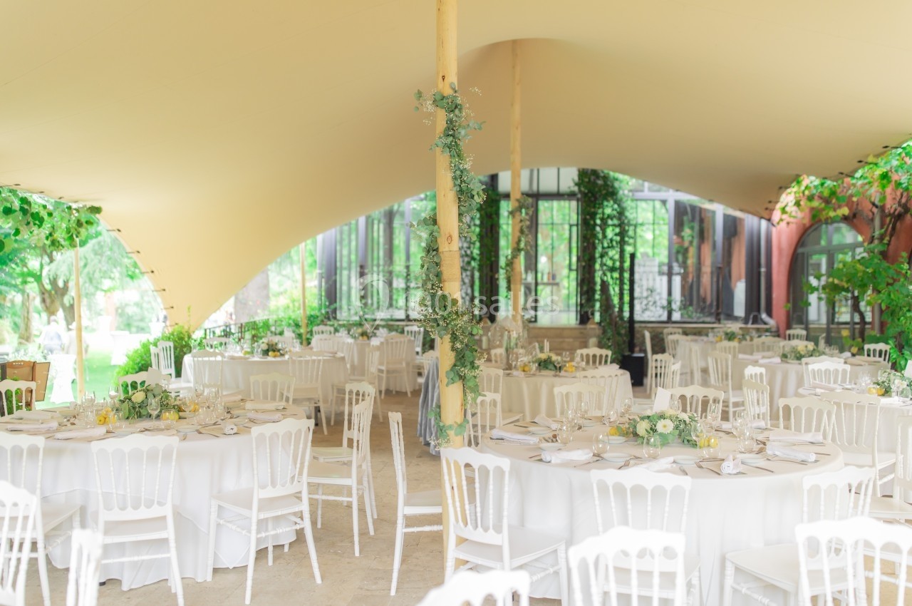 Salle de réception décorée avec des tables rondes blanches, des chaises assorties et des arrangements floraux sous une tente.