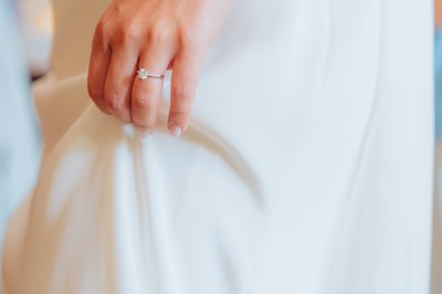 Main d'une personne tenant légèrement une robe blanche, avec un bracelet fin et une bague visible.