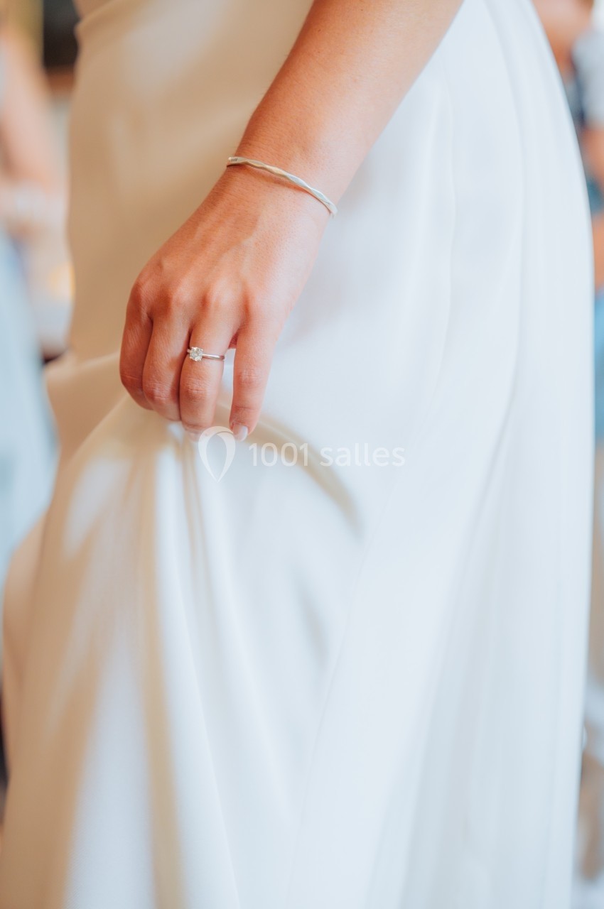 Main d'une personne tenant le tissu d'une robe blanche, avec un bracelet fin et une bague visible.