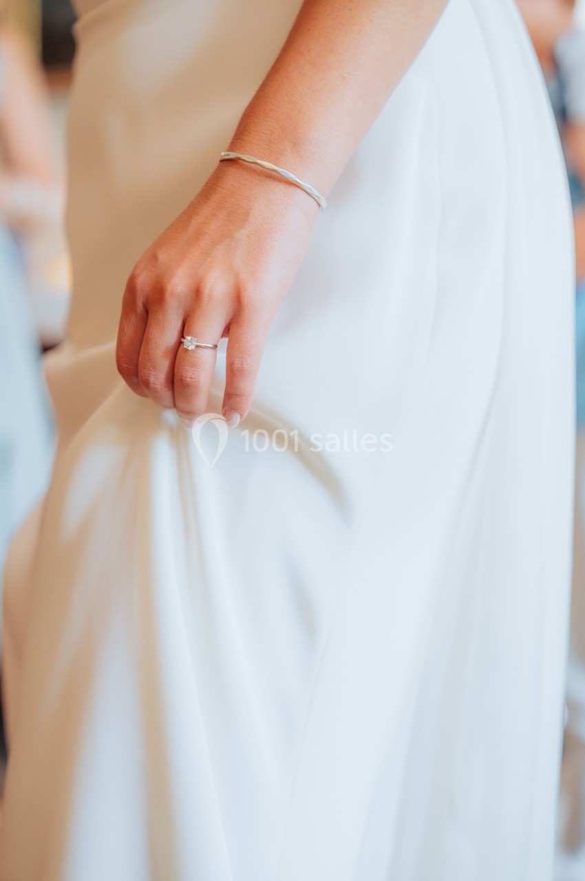 Main d'une personne tenant légèrement une robe blanche, avec un bracelet fin et une bague visible.