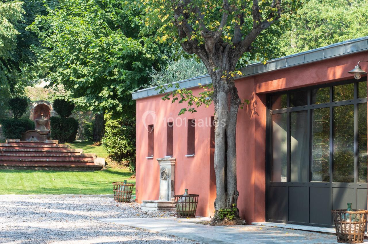 Bâtiment aux murs rouges avec grandes fenêtres, entouré d'arbres et d'une fontaine dans un jardin verdoyant.