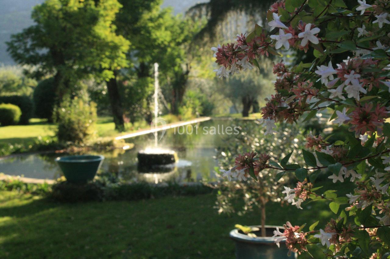 Jardin avec bassin, fontaine, arbres et fleurs en premier plan sous une lumière naturelle.
