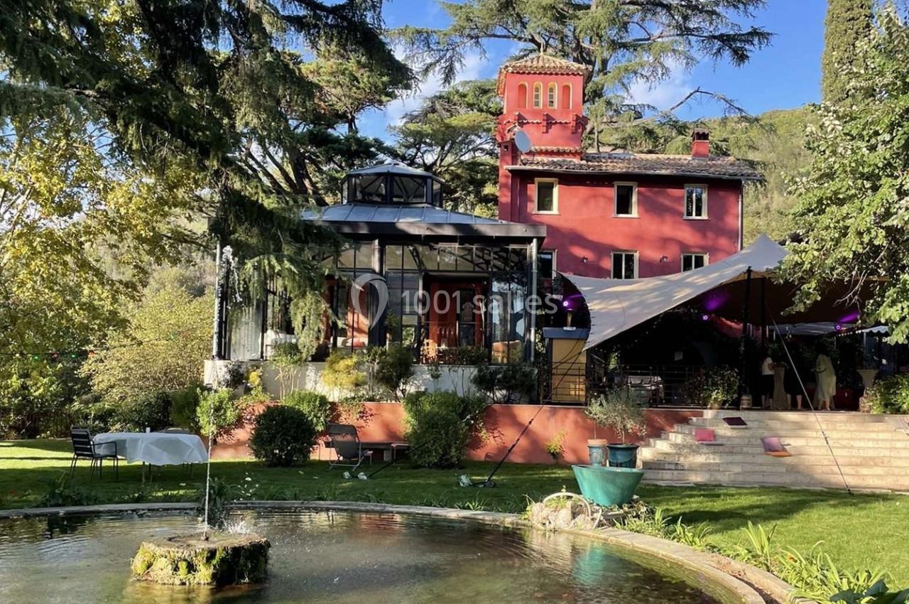 Grande maison rouge avec verrière, entourée d'arbres et d'un jardin avec une fontaine au premier plan.