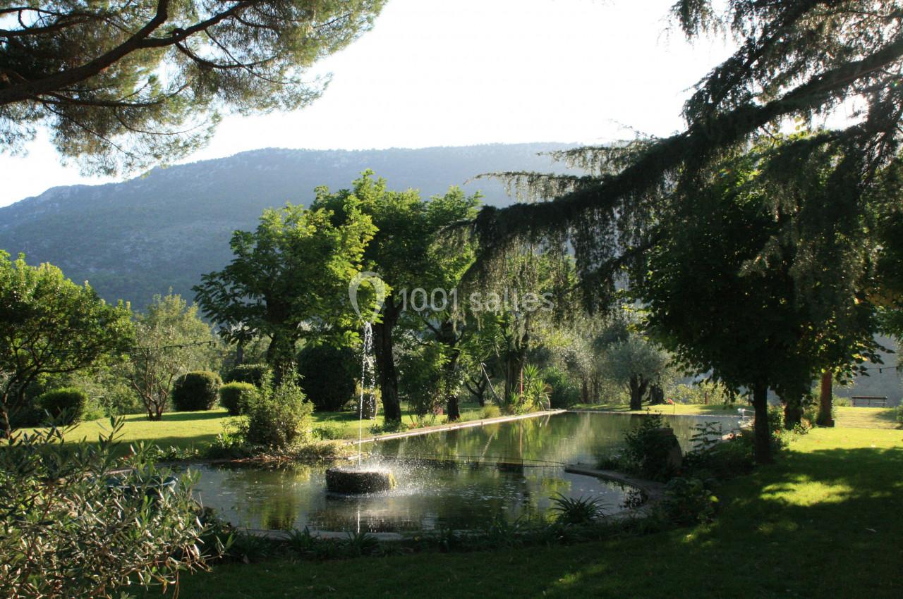 Fontaine entourée de végétation dans un parc verdoyant avec des arbres et une montagne en arrière-plan.