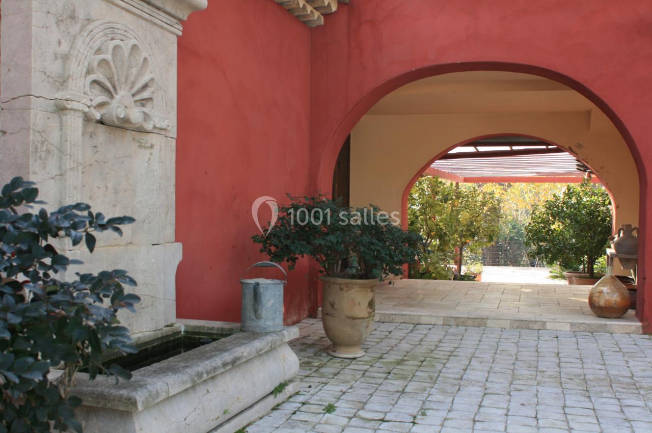 Cour intérieure avec mur rouge, fontaine en pierre, plantes en pot et vue sur un jardin ensoleillé à travers une arche.