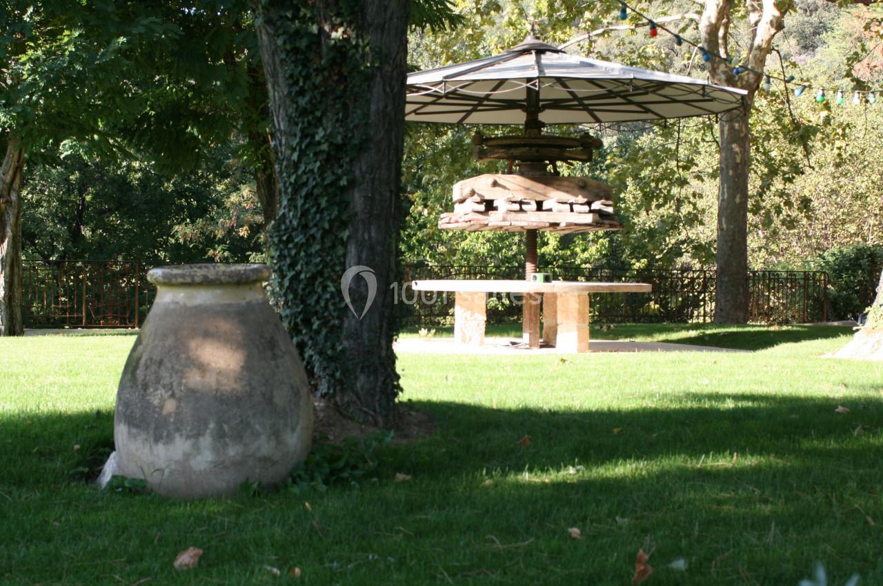 Pot en terre cuite posé sur une pelouse, avec un arbre, une table en pierre et un parasol en arrière-plan.