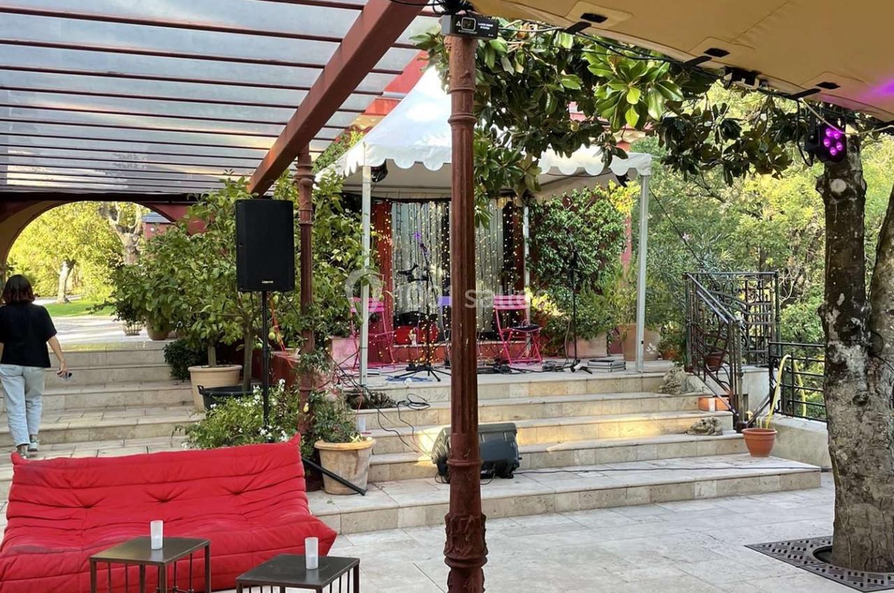 Terrasse extérieure avec canapés rouges, tables basses, scène équipée et végétation environnante sous une pergola.