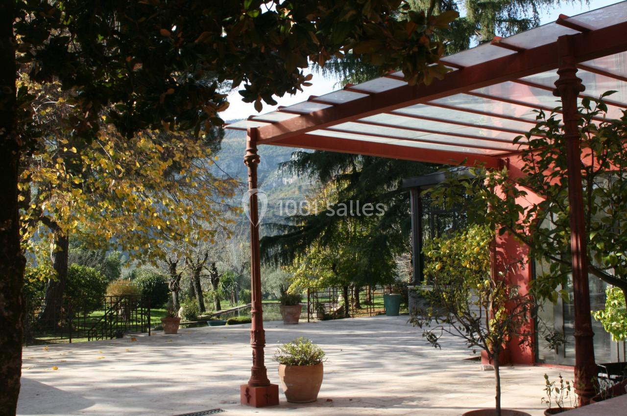 Terrasse couverte par une structure en verre et métal rouge, entourée d'arbres et de pots de fleurs dans un jardin.