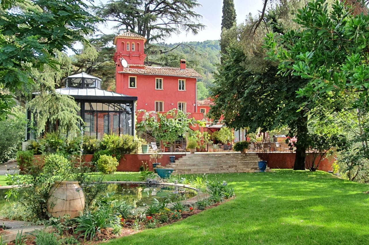Maison rouge entourée de verdure, avec un jardin paysager, un bassin et des arbres, sous un ciel dégagé.