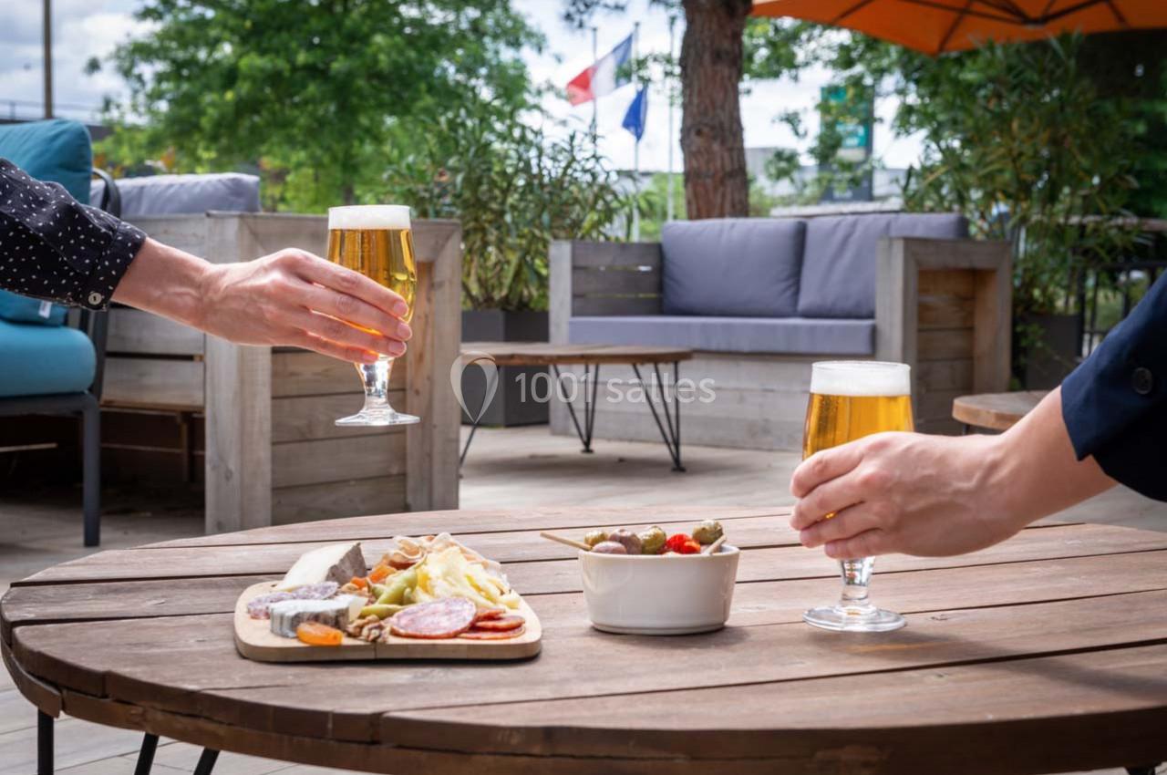 Deux personnes trinquent avec des verres de bière sur une terrasse, près d'une table garnie de charcuterie et d'olives.