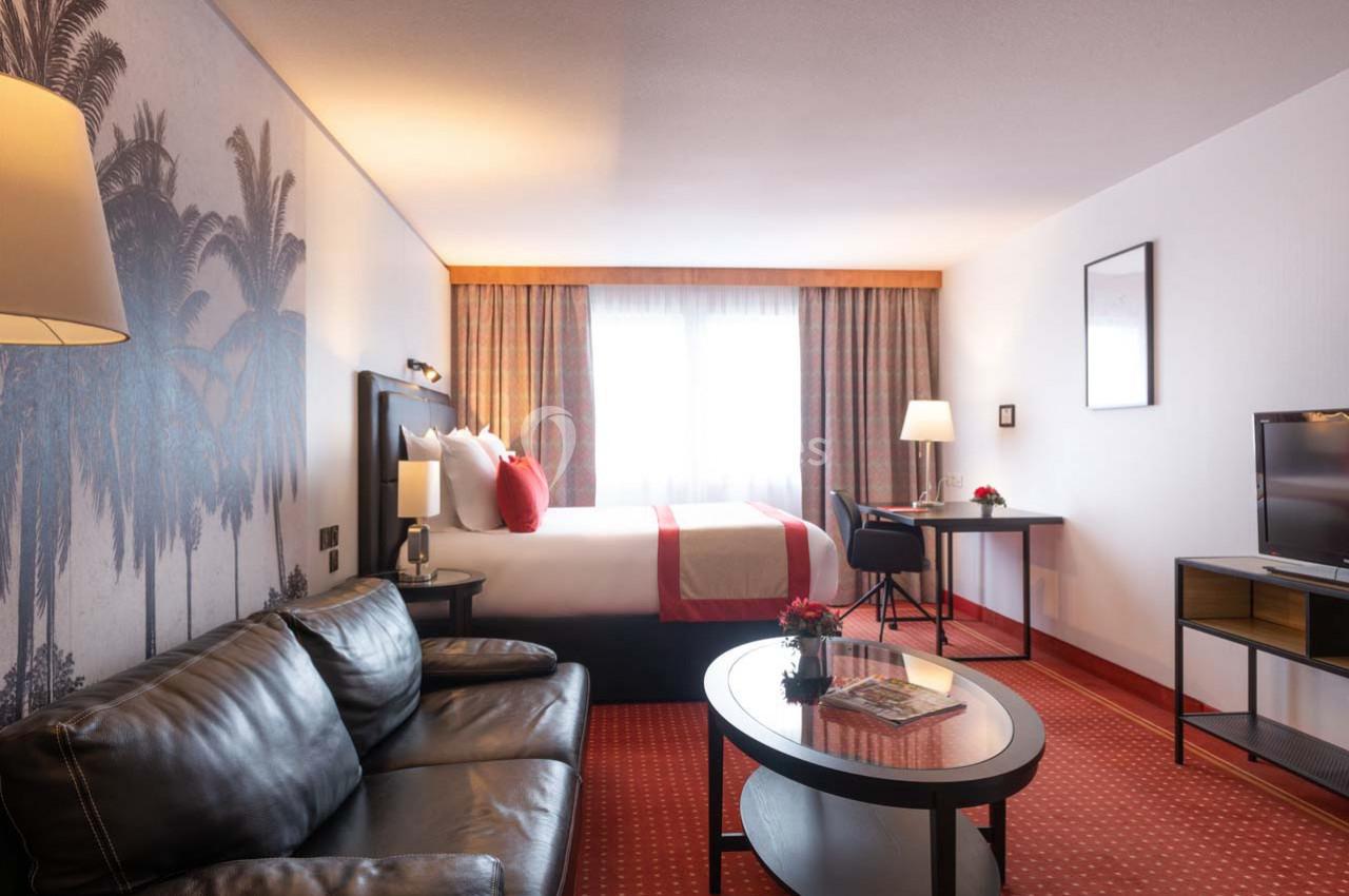 Chambre d'hôtel lumineuse avec lit double, canapé en cuir, bureau, téléviseur et décoration murale de palmiers.