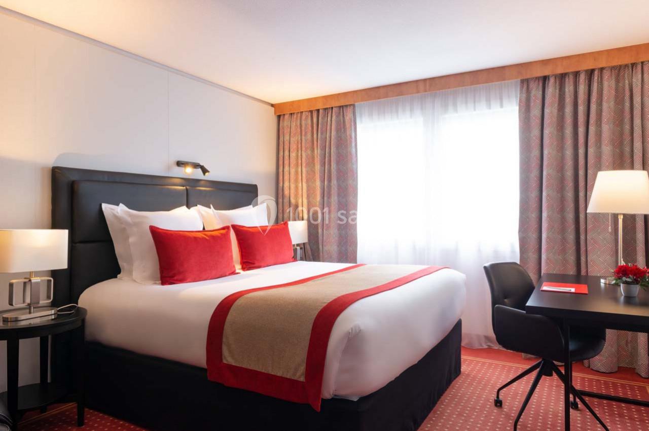 Chambre d'hôtel lumineuse avec un lit double, des coussins rouges, un bureau noir et des rideaux aux motifs discrets.