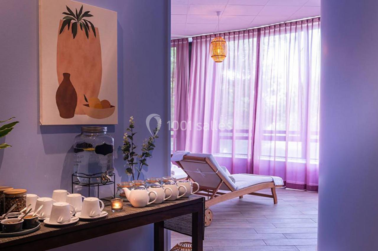 Espace lumineux avec chaises longues, rideaux violets et table garnie de tasses, carafe et plantes décoratives.