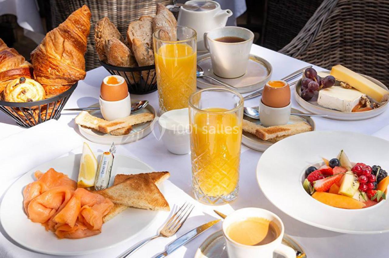 Petit-déjeuner servi sur une table avec viennoiseries, pain, saumon, fruits, œufs à la coque, jus d’orange et café.