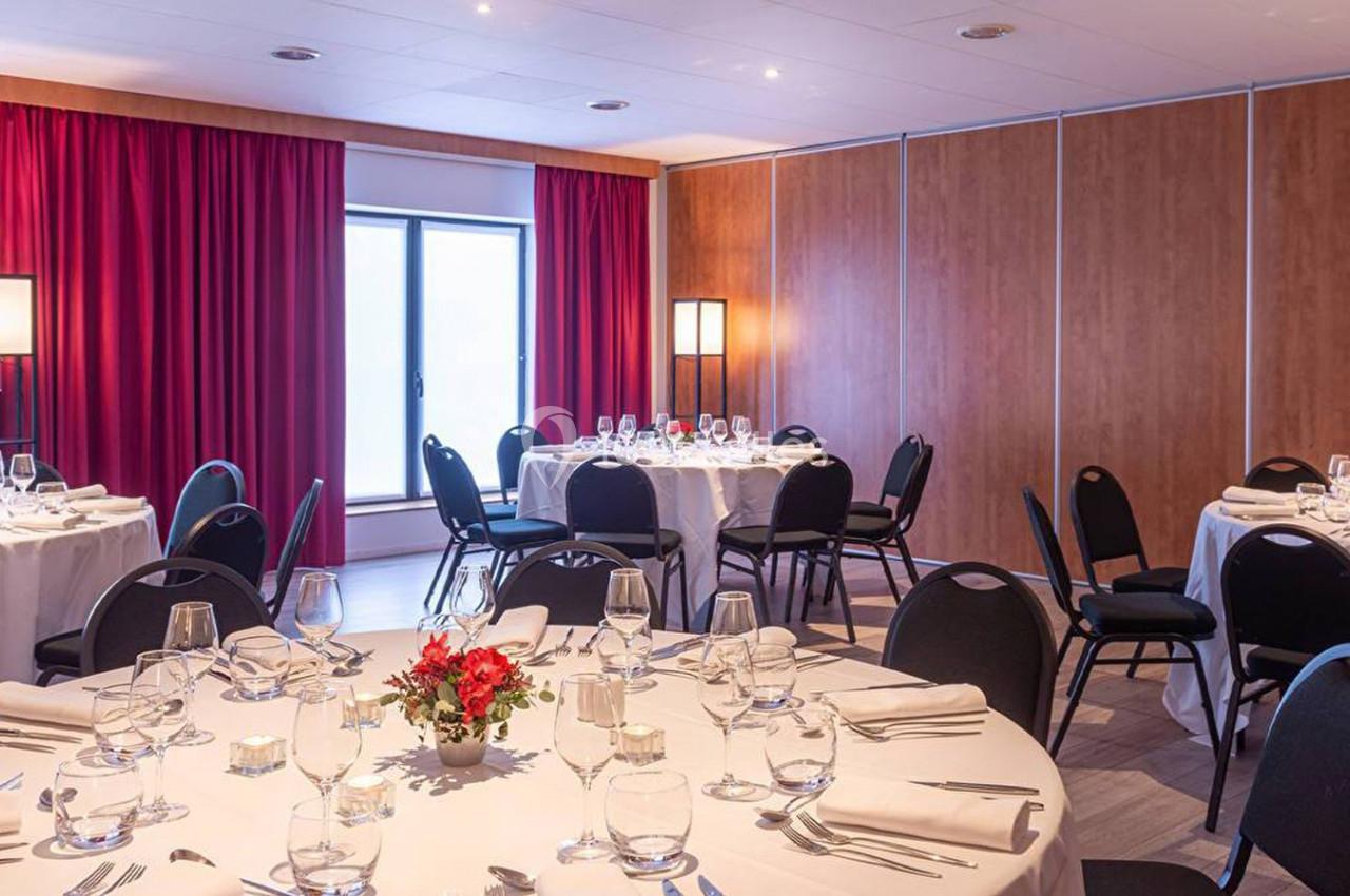 Salle de réception avec tables rondes dressées, nappes blanches, chaises noires et rideaux rouges en arrière-plan.