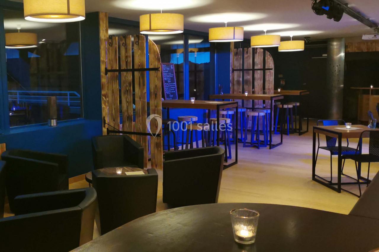 Salle de bar moderne avec éclairage tamisé, tables hautes, tabourets en bois et fauteuils noirs.