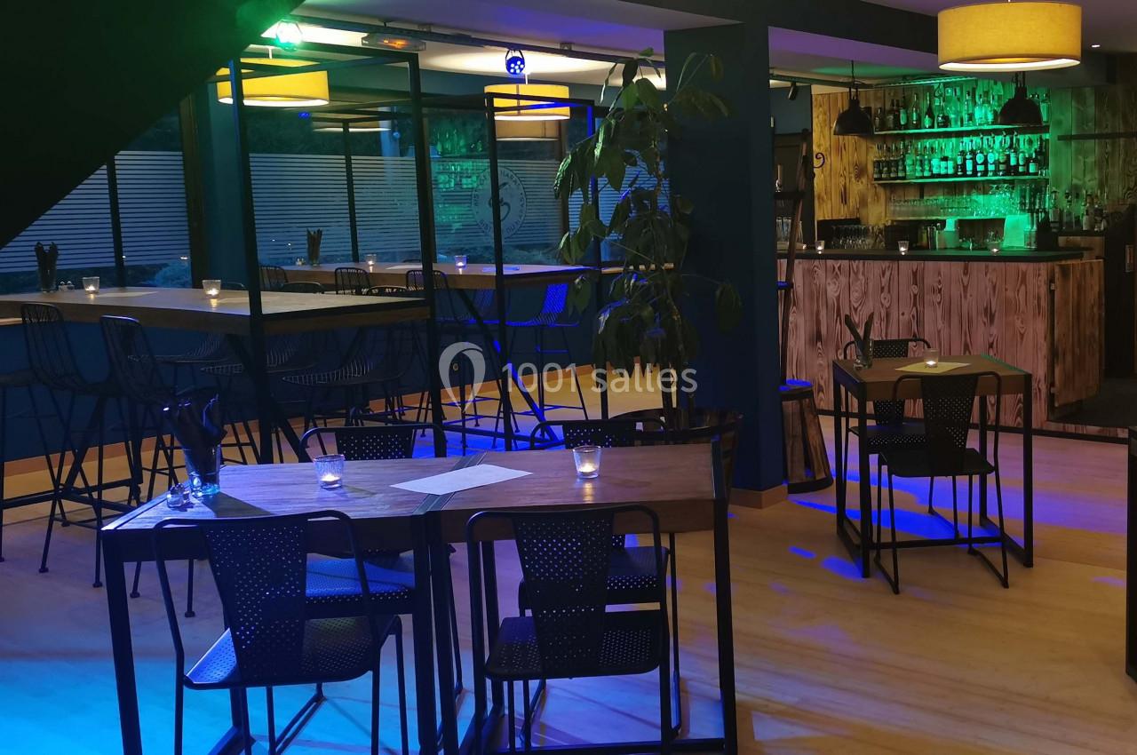 Salle de restaurant moderne avec tables en bois, chaises noires, éclairage tamisé et bar en arrière-plan.