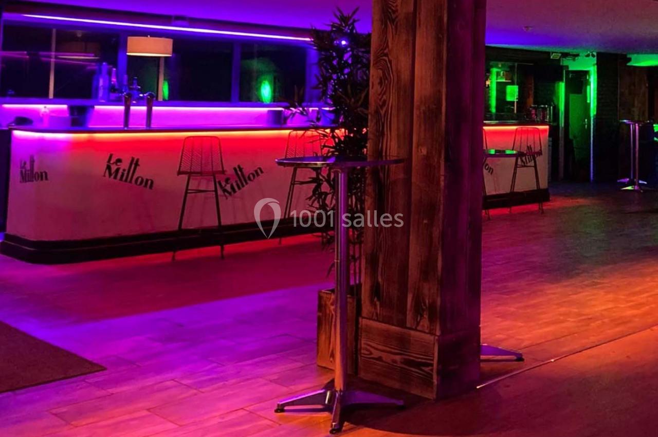 Salle de bar avec comptoirs éclairés par des lumières néon violettes et vertes, sol en bois et tables hautes.