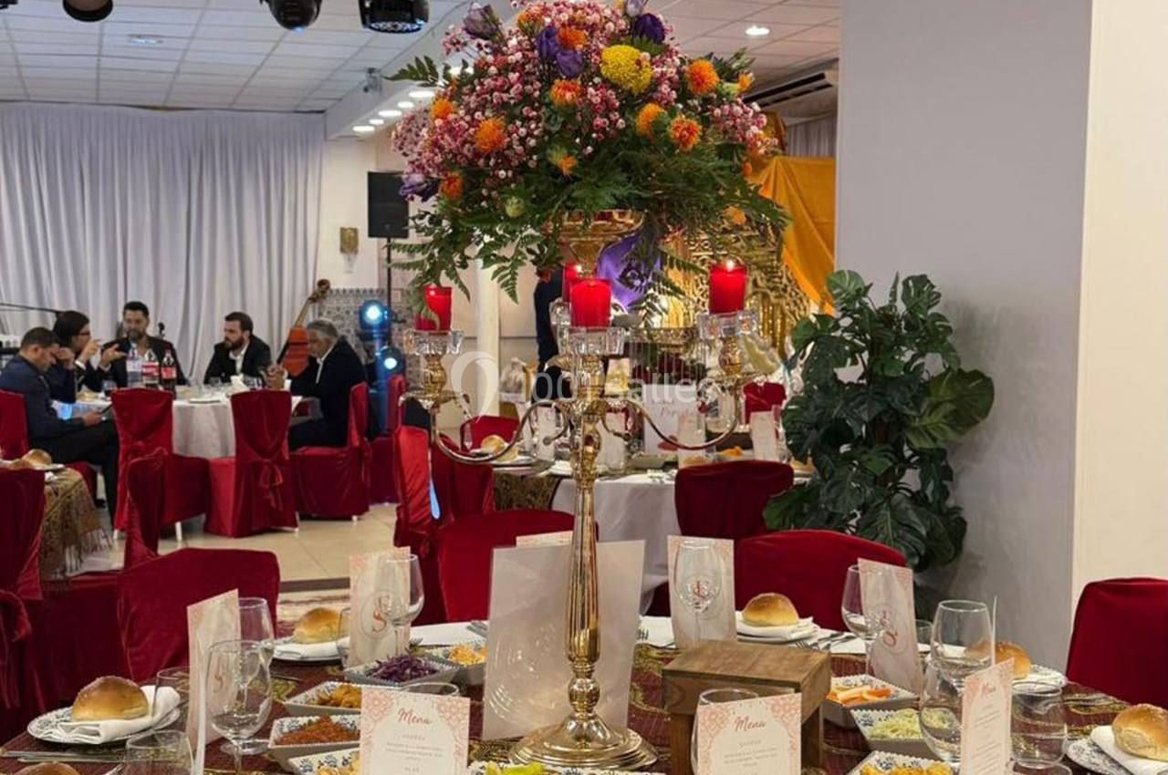 Centre de table avec chandelier doré, fleurs colorées et bougies rouges, entouré de tables dressées pour un événement.