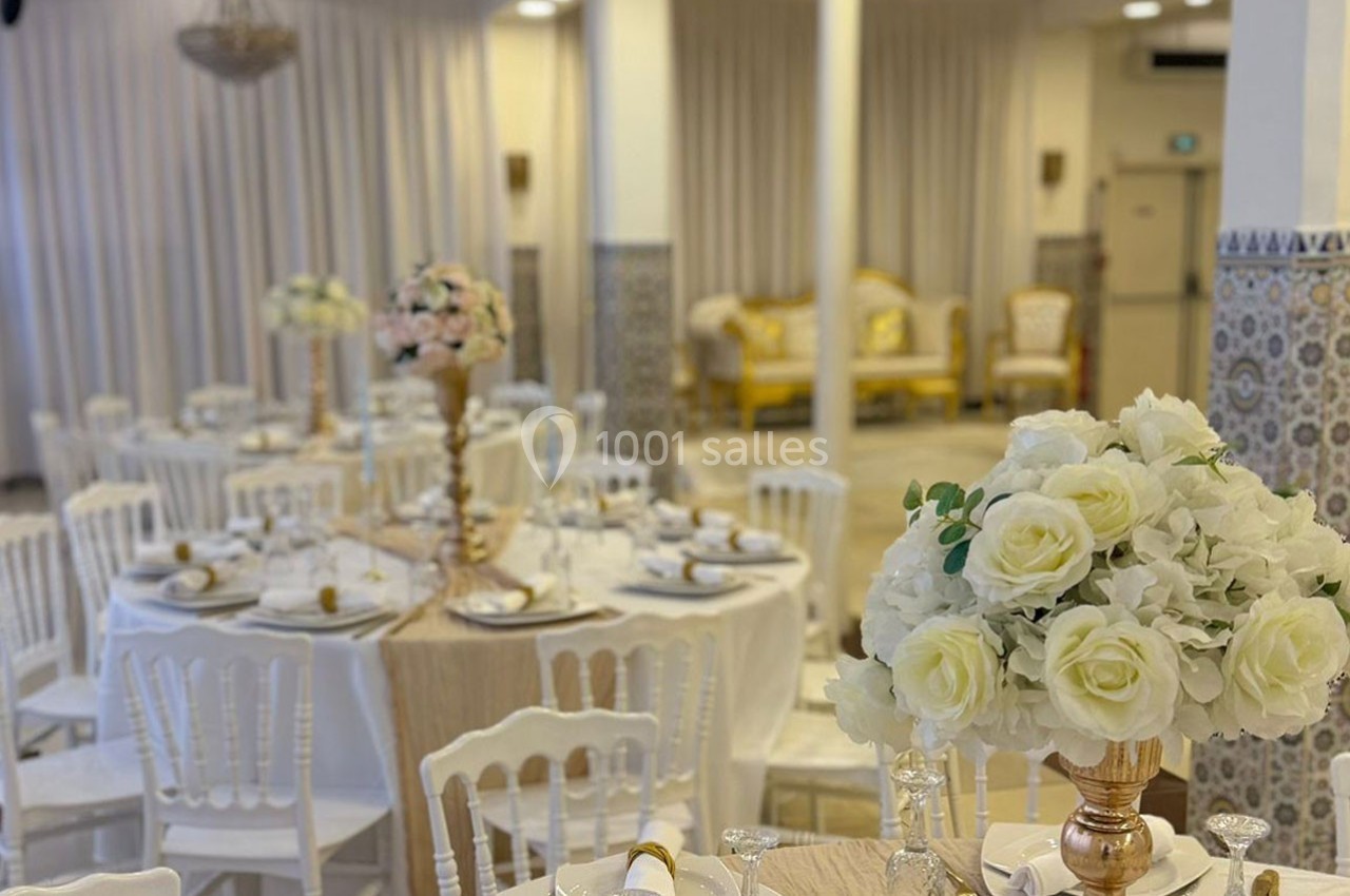 Salle de réception décorée avec des tables rondes, nappes blanches, chaises assorties et centres de table fleuris.