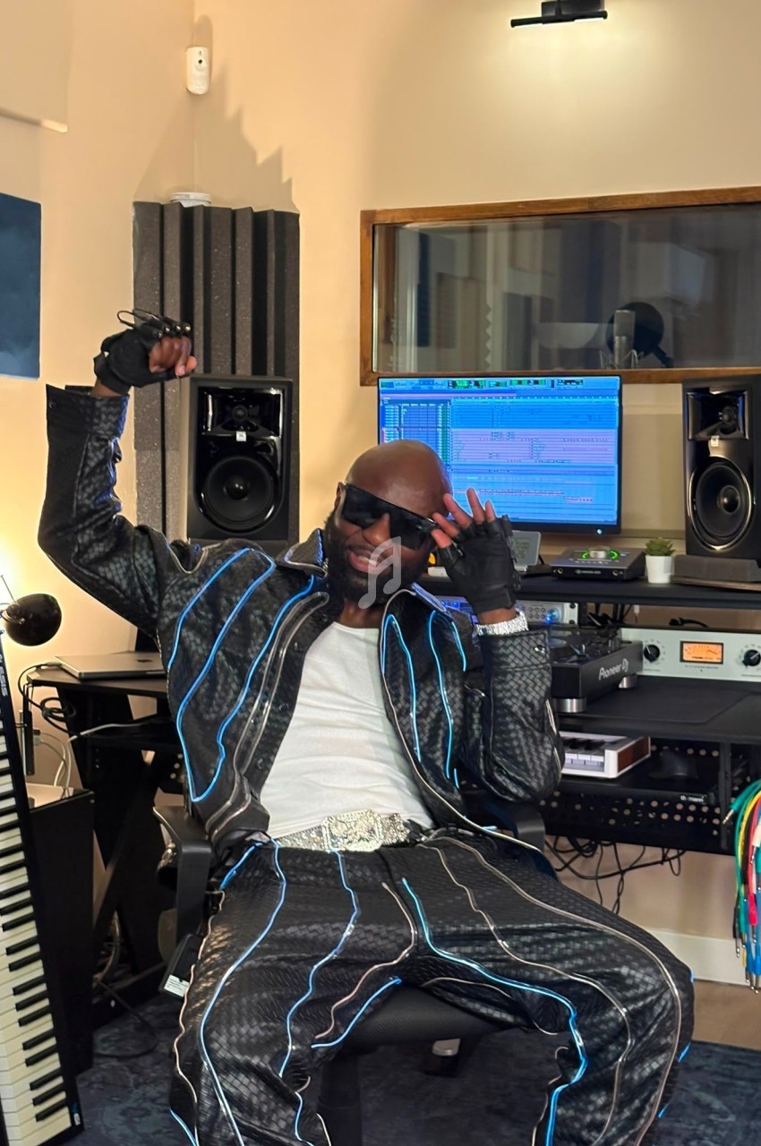 Homme assis dans un studio d'enregistrement, portant une tenue lumineuse, avec des équipements audio en arrière-plan.