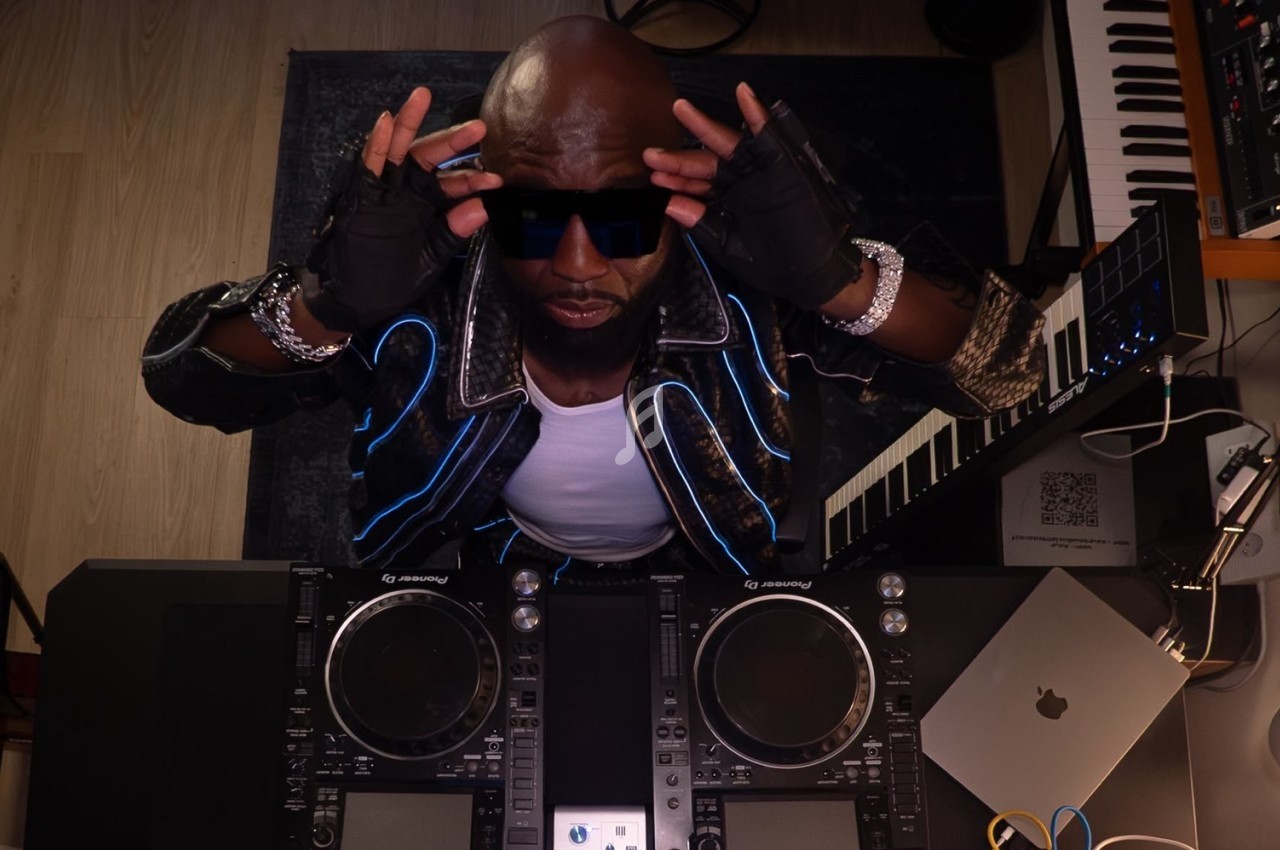 Un DJ portant des lunettes de soleil ajuste sa tenue devant une table de mixage, entouré d'équipements musicaux.
