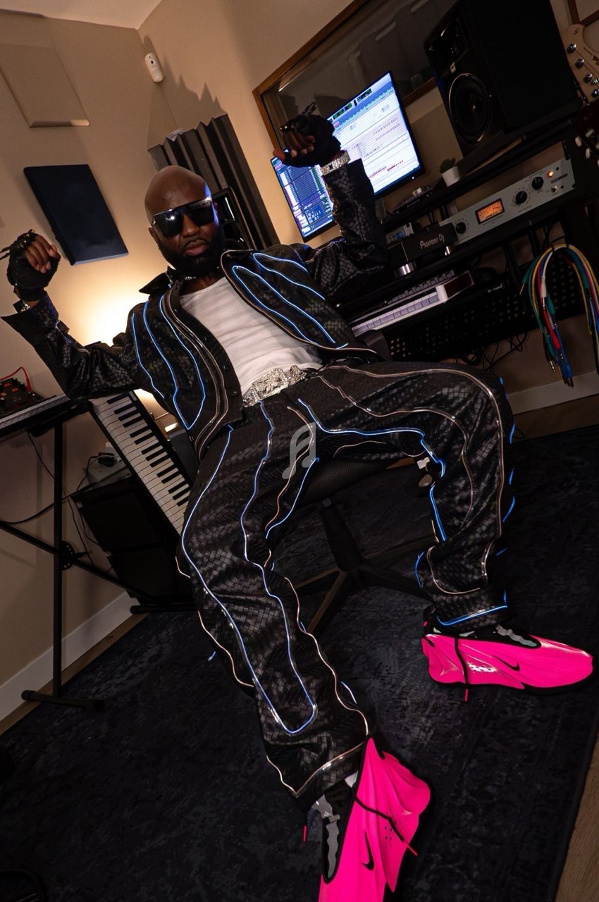 Un homme assis dans un studio d'enregistrement, portant une tenue noire avec des motifs lumineux et des chaussures roses.