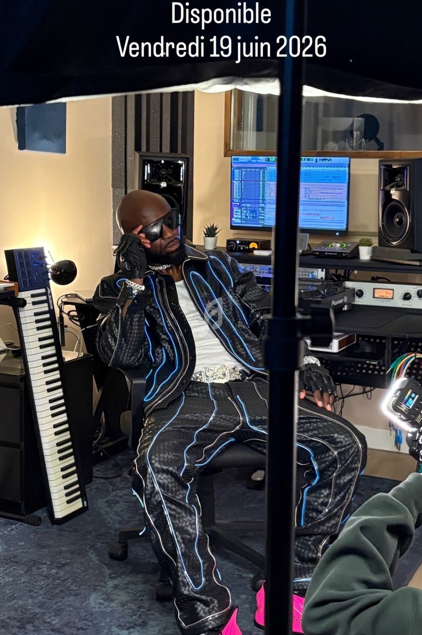Un artiste en studio d'enregistrement, vêtu de vêtements stylés, assis près d'un clavier et d'équipements audio.