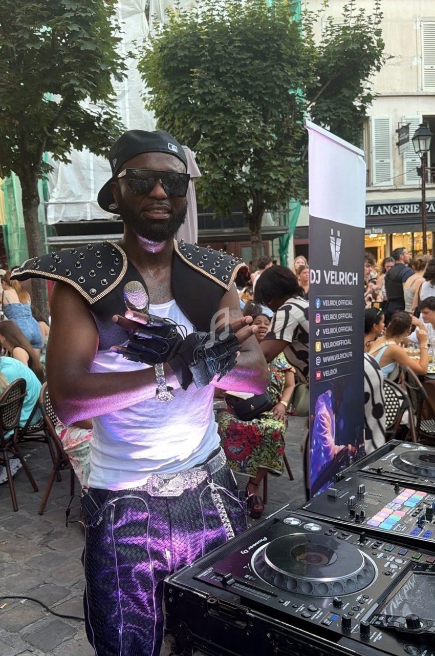 Un DJ en tenue originale mixe en extérieur devant une foule assise à des tables de café en terrasse.