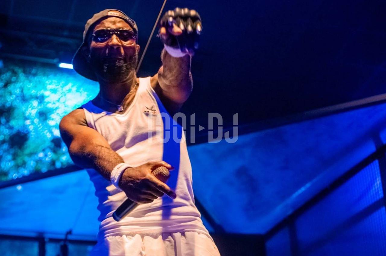 Un chanteur sur scène, tenant un micro, vêtu d'un débardeur blanc et d'une casquette, éclairé par des lumières bleues.