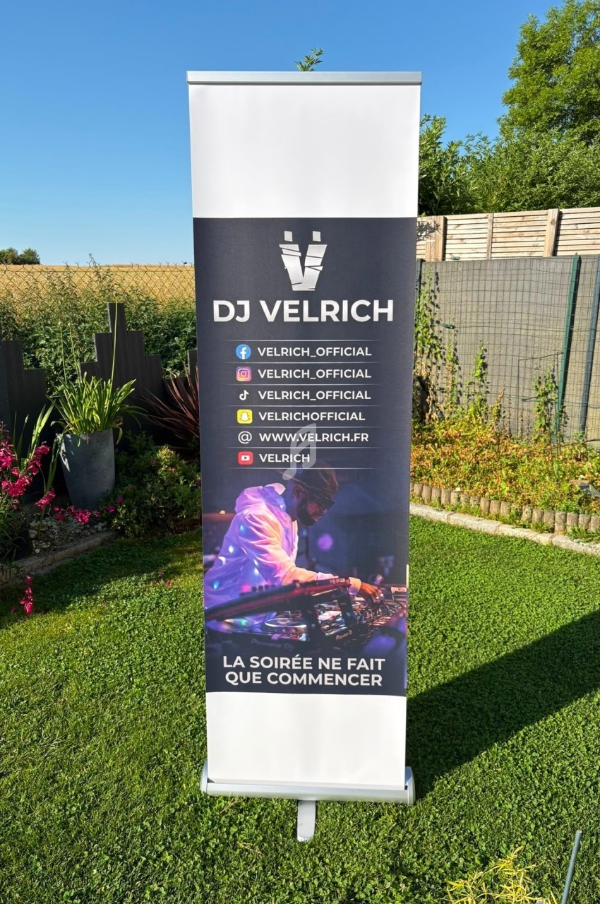 Roll-up publicitaire avec le logo ’DJ Velrich’, des réseaux sociaux et un DJ mixant, installé dans un jardin ensoleillé.