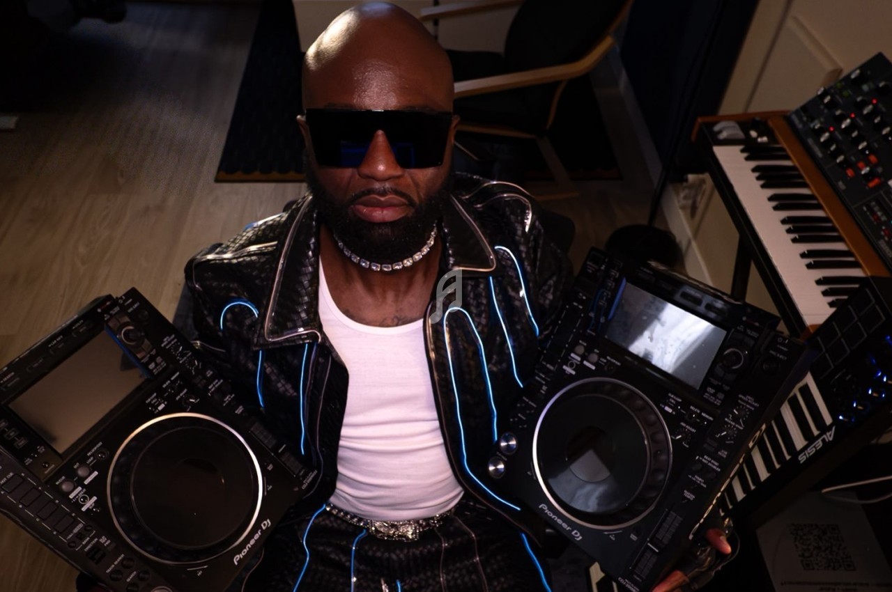 Un homme portant des lunettes de soleil tient deux platines DJ dans un studio équipé d'instruments de musique.