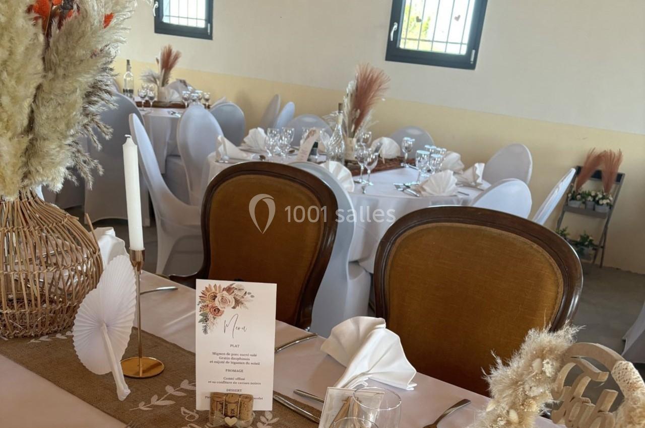 Salle décorée pour un événement, avec tables dressées, chaises blanches, menus et ornements floraux naturels.