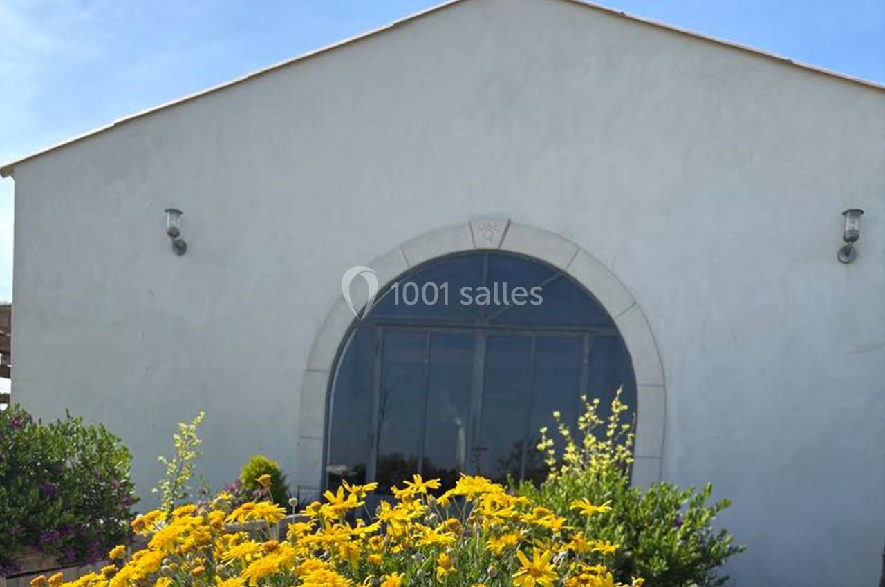 Façade blanche d'un bâtiment avec une grande porte vitrée en arc, entourée de fleurs jaunes et de verdure.
