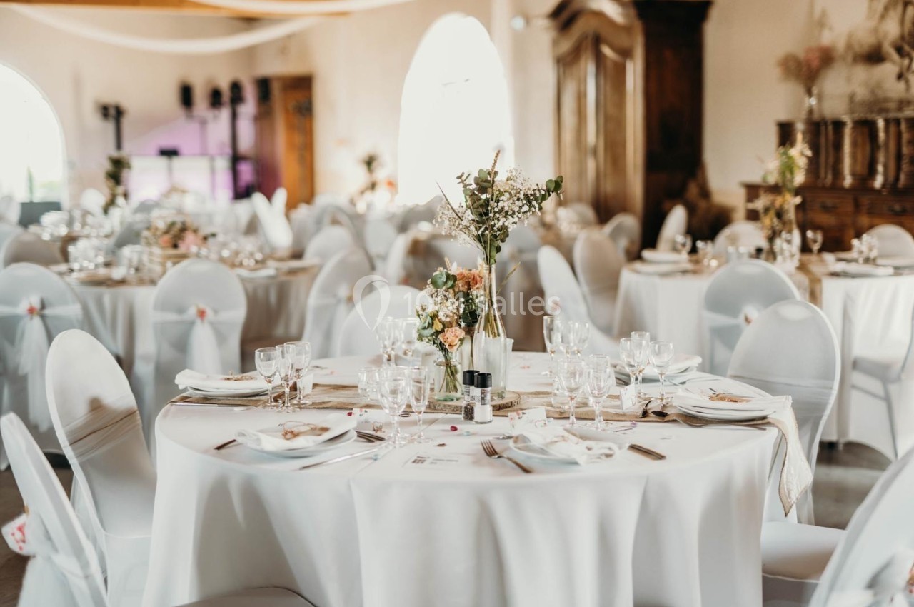 Salle de réception décorée avec des tables rondes, nappes blanches, chaises houssées et centre de table floral.