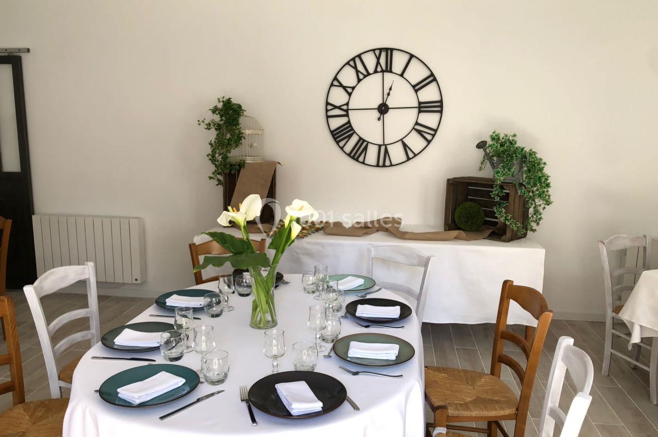 Salle décorée avec une table ronde dressée, des chaises en bois et une grande horloge murale en arrière-plan.