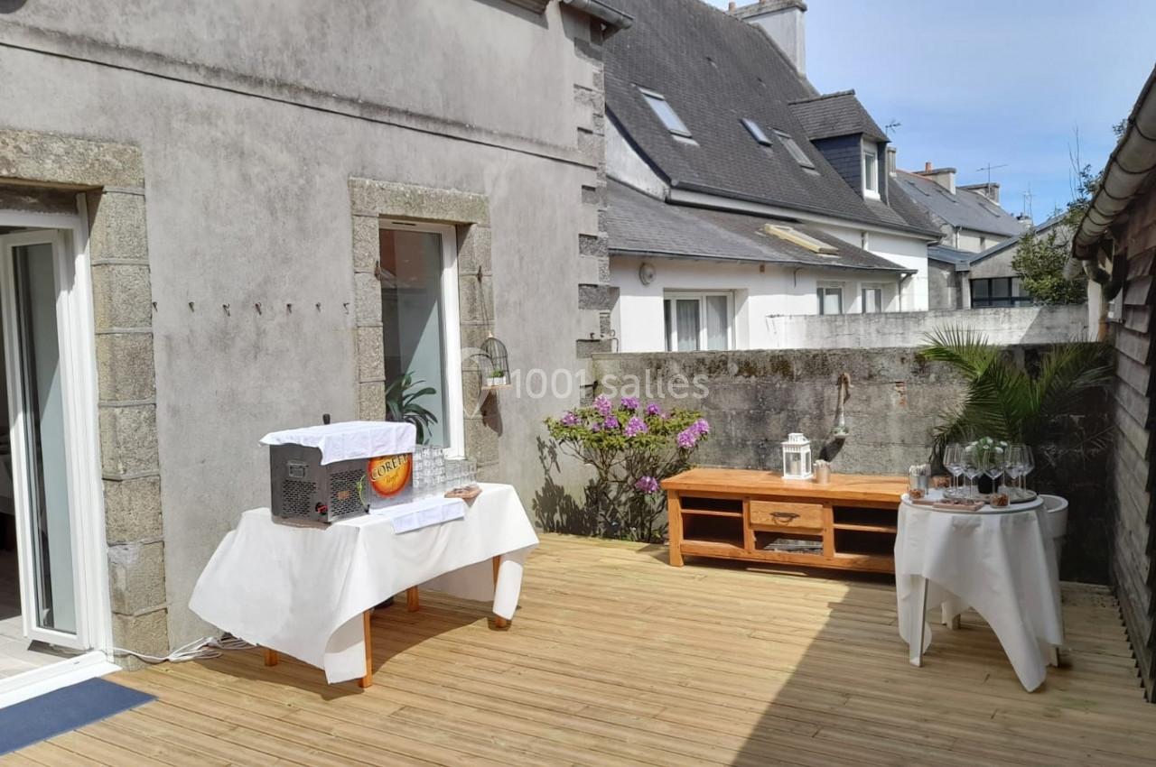 Terrasse en bois aménagée avec des tables décorées, entourée de murs en pierre et de maisons voisines.